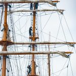 Crew members climbing the tall masts of a vintage sailing ship, surrounded by ropes.
